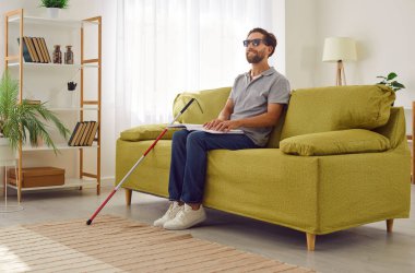 Siyah gözlüklü, sakallı, genç bir adamın portresi. Oturma odasındaki kanepede oturuyor. Sopa bastonuyla ve dokunsal kabartma kitabı okuyor. Körlük ve eğitim kavramı.