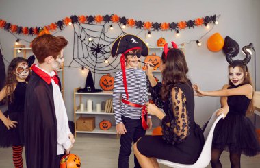 Woman entertainer have fun do makeup for small kids at Halloween celebration at home. Female nanny play with diverse children celebrate all saints eve in costumes in decorated house or kindergarten.