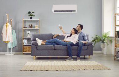 Young family couple enjoying life in their apartment with a modern air conditioning system. Happy husband and wife relaxing on the sofa at home and setting up the temperature on their AC on the wall