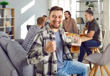 Man having fun and drinking beer at a home party. Portrait of a happy, smiling guy in a plaid shirt holding a beer and looking at the camera while sitting on the couch with friends in the background