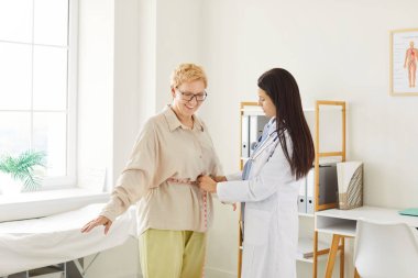 Young doctor, nurse woman holding measurement tape to measure senior patient waist to check after dieting training, fit female in care about body shape, following healthy weight loss, regular exam
