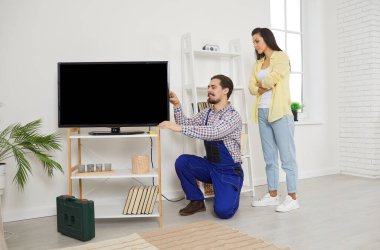 Young friendly service man installs and connects new TV in female clients house. Young woman watches as male repairman in work uniform connects cables to TV screen. Service and maintenance concept.