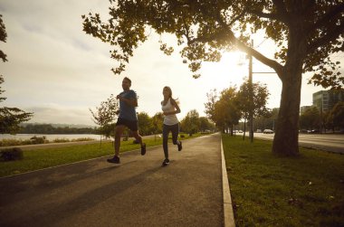 İki kişi şehir sokaklarında aktif koşu antrenmanı yapıyor. Sıcak bir yaz akşamında yeşil ağaçların altında arka planda koşan spor kıyafetli mutlu genç adam ve kadın. Spor konsepti