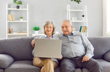 Elderly gray-haired senior couple using laptop having video call and smiling sitting on couch in the living room at home. E-learning, retirement and communication technology concept.