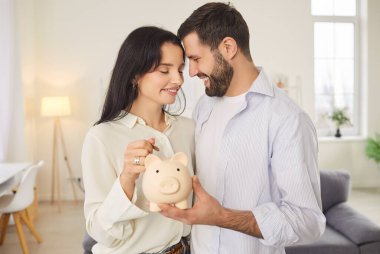 Savings for future, dream or new home. Young couple with piggy bank in their hands radiate happiness as they save money for their future. Man holds piggy bank while woman places shiny coin inside.