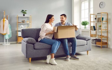 Excited young couple looking at each other while unpacking cardboard box at home. Man and woman customers unboxing parcel receiving online shop order or gift. Delivery and postal service concept