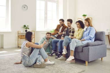 Group of friends hanging out together. Several young people meeting at somebodys place. Young men and women sitting on sofa and listening to girl who is sitting on floor and talking about something
