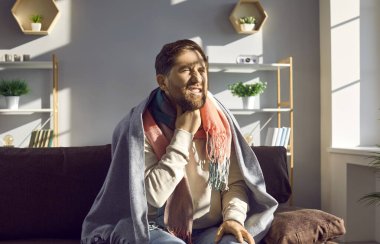 Unhealthy young man feels severe pain and sore throat, malaise and weakness. Bearded man is sitting at home in living room, wrapped in warm blanket, dressed in sweater and jeans. Healthcare concept.