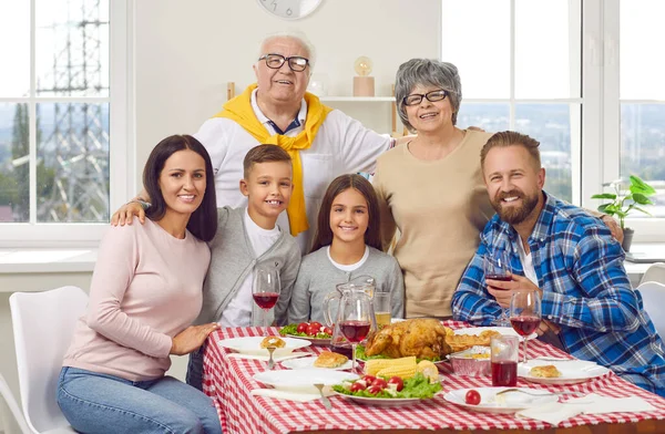 Portrait of happy family of three generations who celebrate Thanksgiving together. Grandparents, young couple and their son and daughter hug and smile together while sitting at festive table at home.