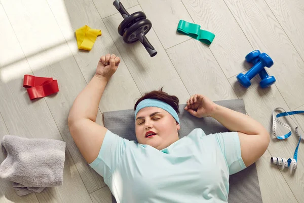 Portrait of a funny young fat overweight woman tired after fit sport exercises wearing sportswear lying on the floor at home or gym and having a rest. Workout, fitness and body positive concept.