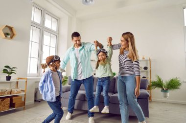 Carefree cheerful parents, son and daughter playing at home. Happy mother and father having fun carrying their daughter wearing goggles by hands like airplane in living room