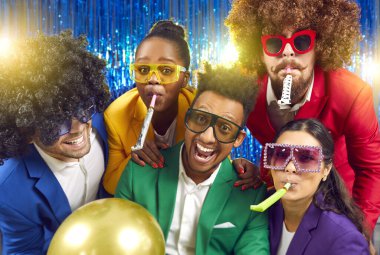 Portrait of funny young people in different party glasses, wigs and with festive whistles having fun together at party. Multiracial friends in colorful clothes laughing against blue shiny foil curtain