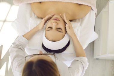 Cosmetologist at spa salon doing facial massage for young woman. Beautiful young girl relaxing and enjoying professional beauty treatment that helps with tension relief and lymphatic drainage