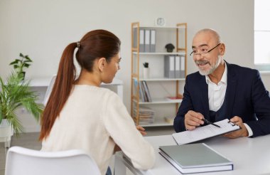 Senior manager talking to a client in the office. Young woman listening to a friendly bank worker, loan broker, realtor or insurance agent whos offering her a deal and showing the contract agreement
