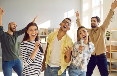 Happy multiracial friends have fun sing karaoke together at home. Smiling millennial diverse multiethnic young people enjoy leisure weekend activity. Hobby and entertainment. Technology concept.