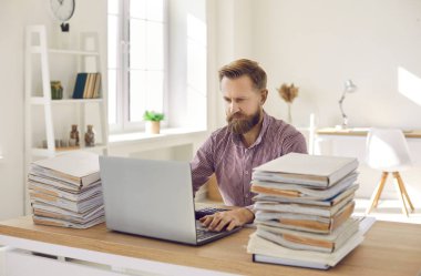 Serious man working in the office. Busy employee doing reports. Focused financial accountant sitting at his desk with lots of paperwork and working on his laptop computer