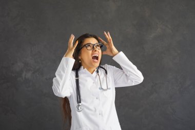 Angry mad stressed tired doctor in white lab coat with stethoscope standing against grey office or studio wall shouting fed up with endless work hours, night shifts, stupid patients and boss orders