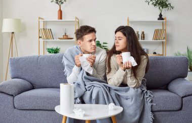 Young unhealthy couple sitting on sofa in the living room covered with plaid, suffering from flu or cold. Sick man and woman looking at each other, holding cups in their hands and drinking hot tea.