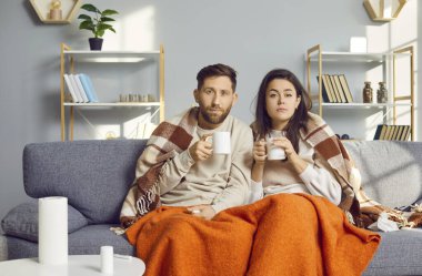 Young couple covered in blankets sitting on sofa at home with seasonal flu or cold. Sick man and woman looking at camera and drinking hot cup of tea beverage feeling unhealthy with influenza.