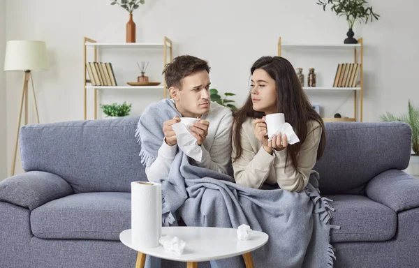 Young unhealthy couple sitting on sofa in the living room covered with plaid, suffering from flu or cold. Sick man and woman looking at each other, holding cups in their hands and drinking hot tea.