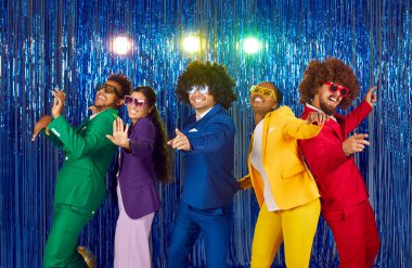Group of funny diverse people in bright costumes dancing at disco party. Five happy multiracial friends in colourful suits, crazy curly hair wigs, funky glasses dancing on blue foil fringe background