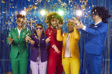 Portrait of funny young people having fun together having fun at New Year themed retro party. Friends in colorful clothes, wigs and glasses laughing and throwing confetti against blue foil curtain.