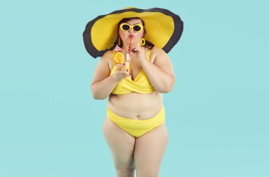 Portrait of fat woman in yellow swimsuit, beach hat and sunglasses drinking orange juice cocktail and looking cheerful at camera on blue background. Summer holiday trip and vacation concept.
