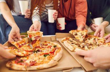 Close up hands of a young people friends gathering in pizzeria or at home together eating tasty Italian food taking pizza slices from box enjoying party with cardboard cups in hands.