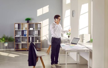 Architect or cadastre engineer talking on phone. Young man standing by office desk with city maps, cadastral plans and laptop, holding sheet of paper and talking on mobile phone