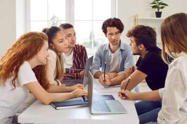 School, college or university students working on group project. Several young people sitting around table with laptop computers, discussing their creative assignment and writing ideas in notebooks