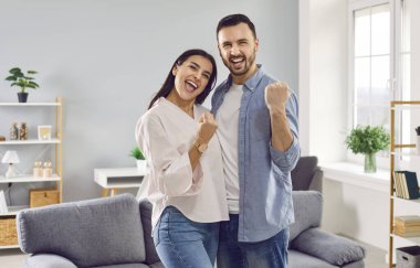 Portrait of a happy excited overjoyed couple family with yes gesture standing in the living room at home looking at the camera like a winners. Buying new apartment,sale or discount offer concept.