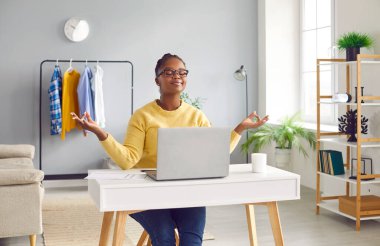 Woman takes break for meditation. Happy beautiful African American girl sitting at her working desk with laptop computer at home, doing mudra gestures, meditating and relaxing. Stress relief concept