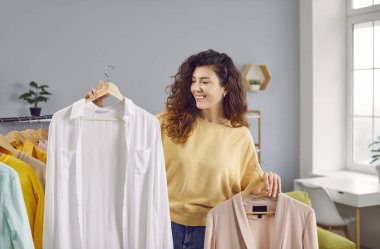 Beautiful woman choosing clothes on rack at home. Attractive smiling young woman holding hangers with shirts, thinking what to wear. Girl trying to choose outfit dressing