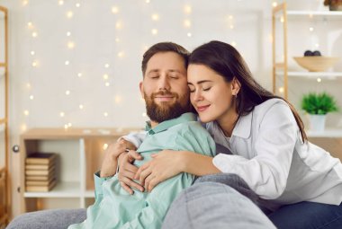Happy couple enjoying time together. Young family spending time at home. Portrait of a beautiful young loving woman standing behind the couch and tenderly hugging her husband