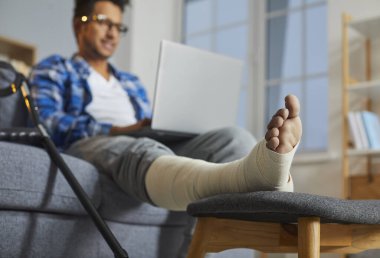 Young man with broken leg sitting on sofa using laptop. Man with plastered limb working, having online meeting, browsing internet at home, selective focus, blurred background