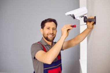 Technician man in uniform installing CCTV camera on wall. Smiling male worker checking and maintaining alarm system on wall using screw driver. Home security system concept