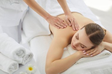 Relaxed woman lying down, massage in spa saloon, cosmetologist hands massaging, body relaxing treatment in beautician room, stack of white towels and asian flower, massage bed covered in white linens