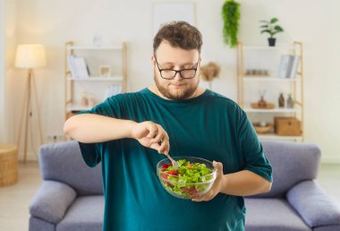 Overweight man eating salad with fork. He mixes a bowl of fresh vegetables at home, embracing healthy diet. Cozy living room background. Healthy lifestyle and mindful eating concept.