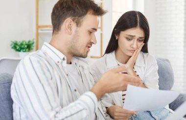 Serious stressed young couple at home worried about paper document, discussing budget, unpaid bank debt, bills, shocked poor family looking counting loan payment upset about money problem, thinking