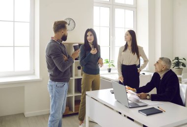 Team of happy corporate employees talking during coffee break at work. Group of four people of different ages gather in their cozy modern office, share news and discuss latest projects together