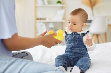 A curious baby sits on a bed in denim overalls while a mother offers a yellow rubber duck, creating a playful, tender moment in a softly lit family bedroom. Baby reaches for the rubber duck.