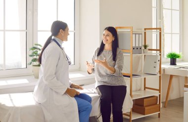 Happy smiling woman patient talking with her friendly young doctor wearing stethoscope holding report file with appointment sitting on the couch in office during medical examination in clinic.