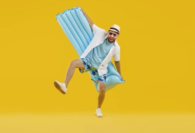 Funny overjoyed excited man wearing beach clothes with open mouth holding inflatable mattress isolated on a studio yellow background. Happy tourist is going on summer holiday trip. Vacation concept.