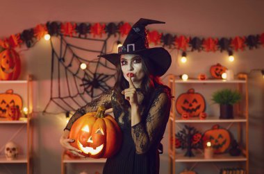 Halloween theme. Portrait of mysterious woman witch making gesture of silence asking not to reveal secrets. Young woman in witchs clothing puts finger to her mouth saying shhh to camera.