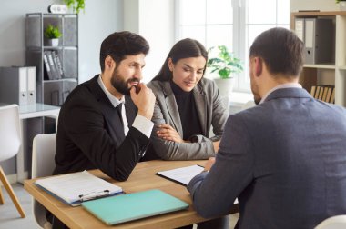 Businesspeople sitting at desk, reviewing contract documents, discussing details, pondering, examining paper, presenting information on business meeting, agent and couple client serious consultation