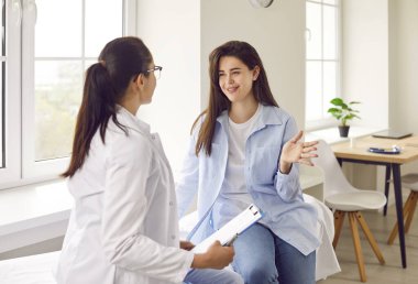Happy smiling woman patient talking with her friendly young female doctor therapist holding report file with appointment sitting on the couch in office during medical examination in clinic.