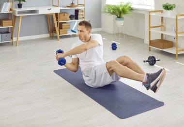 Fit, sporty, young man having a fitness workout at home, sitting on a sports mat, and doing an active physical exercise with dumbbells. Body training, healthy lifestyle concept