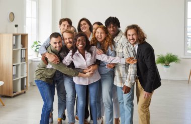 Group portrait of cheerful multiracial colleagues in office hugging to show unity and support at work. Funny diverse men and women in casual clothes hugging and smiling at camera. Team concept.