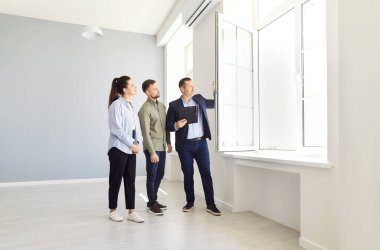 Young couple and agent inspect empty house. Real estate salesman opens window in new apartment. Concept rental housing, real estate agent, final walkthrough, new apartment, people meeting