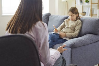 Therapist listening child writing in notebook during counseling session in modern psychologists office. School boy sitting on sofa with pencil to write notes on support consultation with therapist